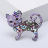 Cat brooch with purple enamel, pink stones, floral details, and green leaf accents, upright pose on white studio backdrop.