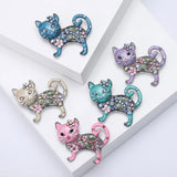 Set of five cat brooches in assorted enamel colors with floral and faceted stone details, arranged on white display blocks.