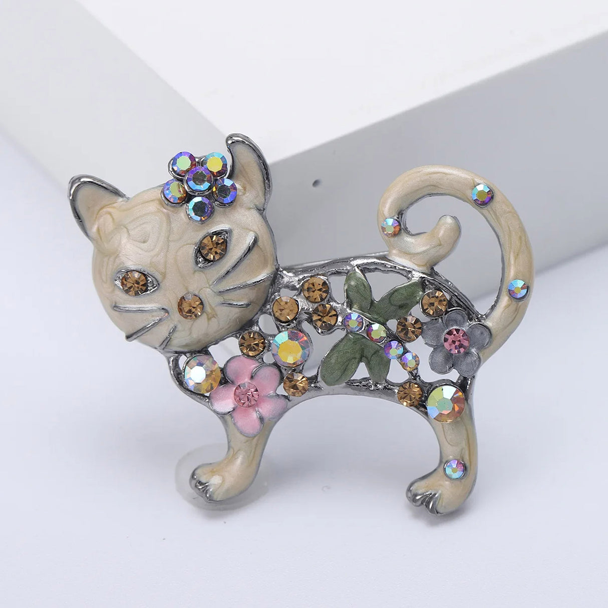 Cat brooch in cream-beige enamel with amber-colored stones, pink flower detail, and green leaf accent, front studio view.