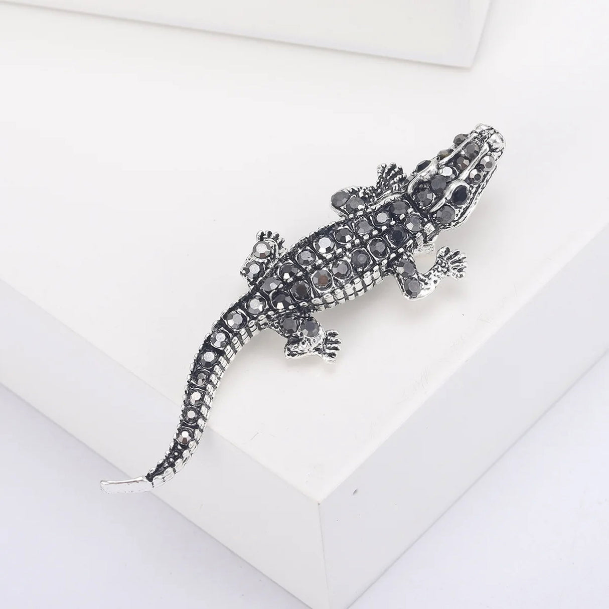 Lizard brooch in silver-tone metal with black faceted stones, angled top-down view on white platform in studio.