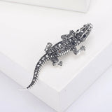 Lizard brooch in silver-tone metal with black faceted stones, angled top-down view on white platform in studio.