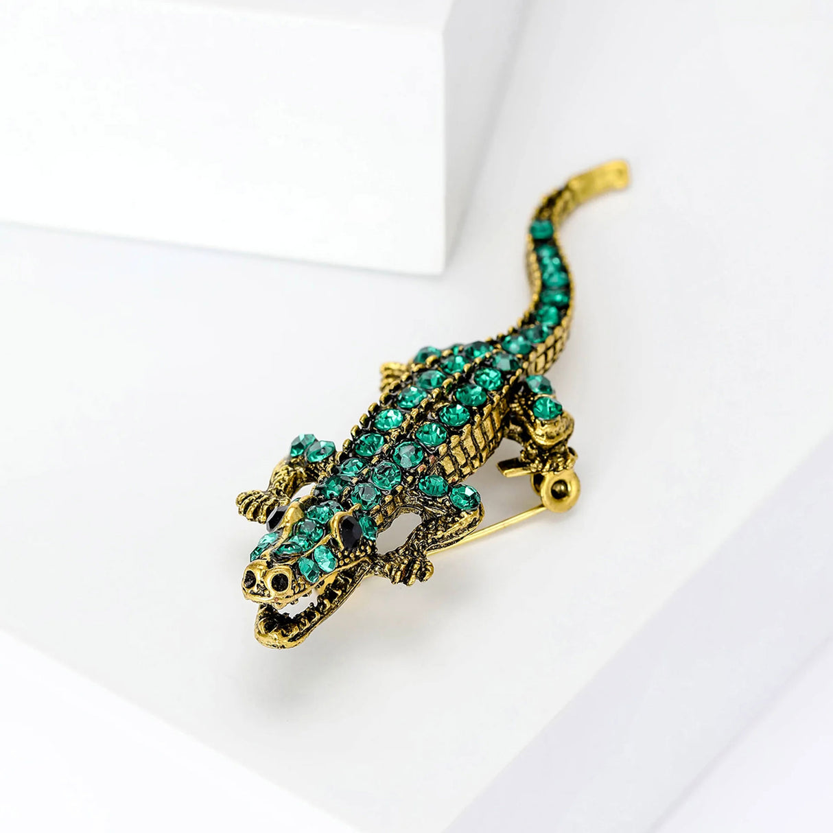 Crocodile brooch in gold-tone with green faceted stones and open jaw detail. Angled front view on white platform in studio setting.