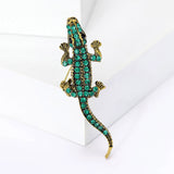 Crocodile brooch with gold-tone metal finish and green faceted stones, upright pose against white studio backdrop.