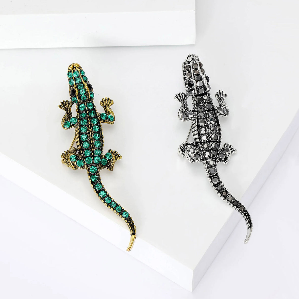 Set of two crocodile brooches, one in gold-tone with green stones and one in silver-tone with black stones, side-by-side on white platform in studio.