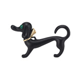 Dachshund dog brooch with matte black finish, green faceted eye, and gold-tone bow accent, cutout on white background.