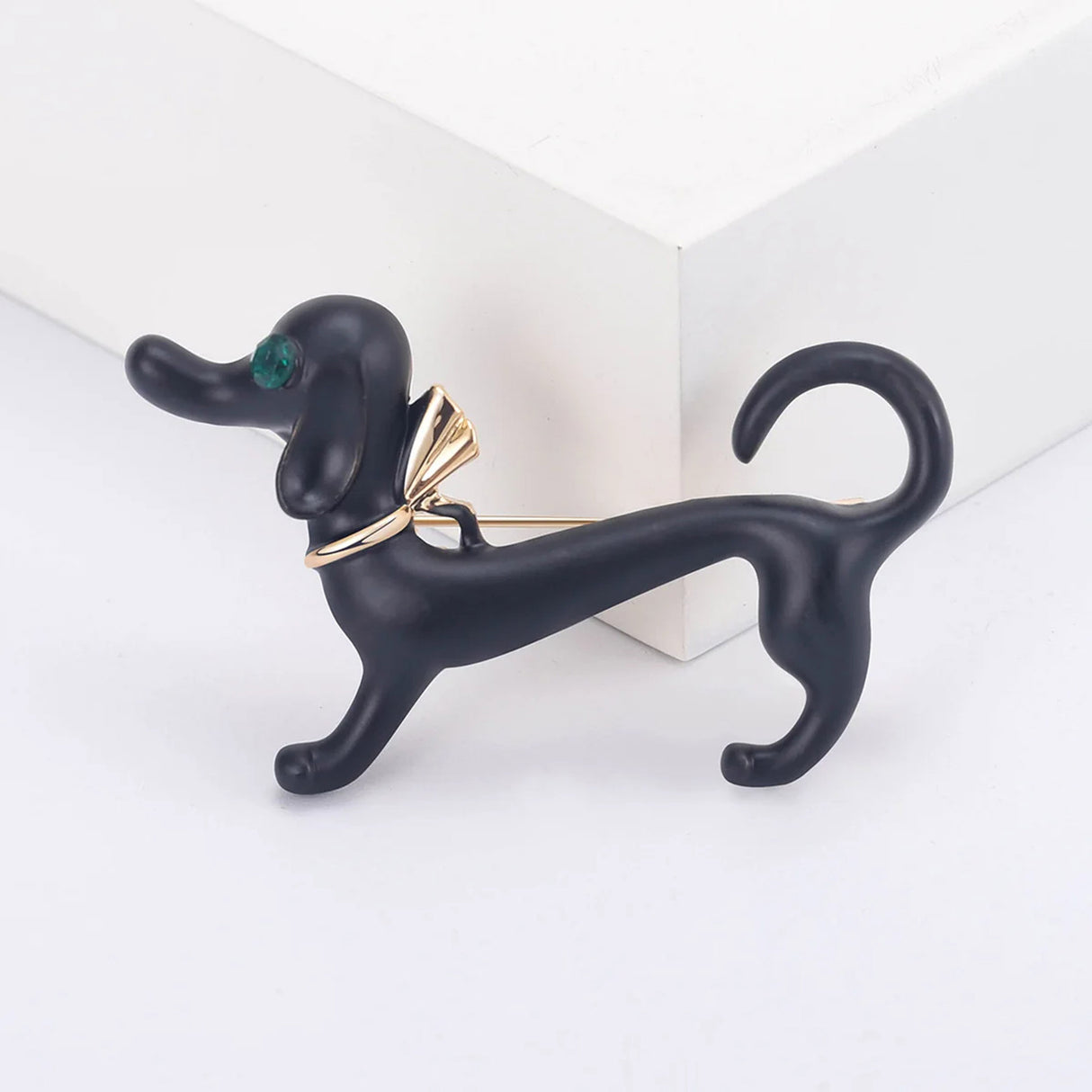 Ddachshund dog brooch in matte black finish with green faceted eye and gold-tone bow detail, displayed upright on white studio surface.