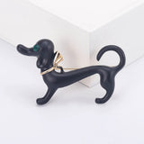 Ddachshund dog brooch in matte black finish with green faceted eye and gold-tone bow detail, displayed upright on white studio surface.