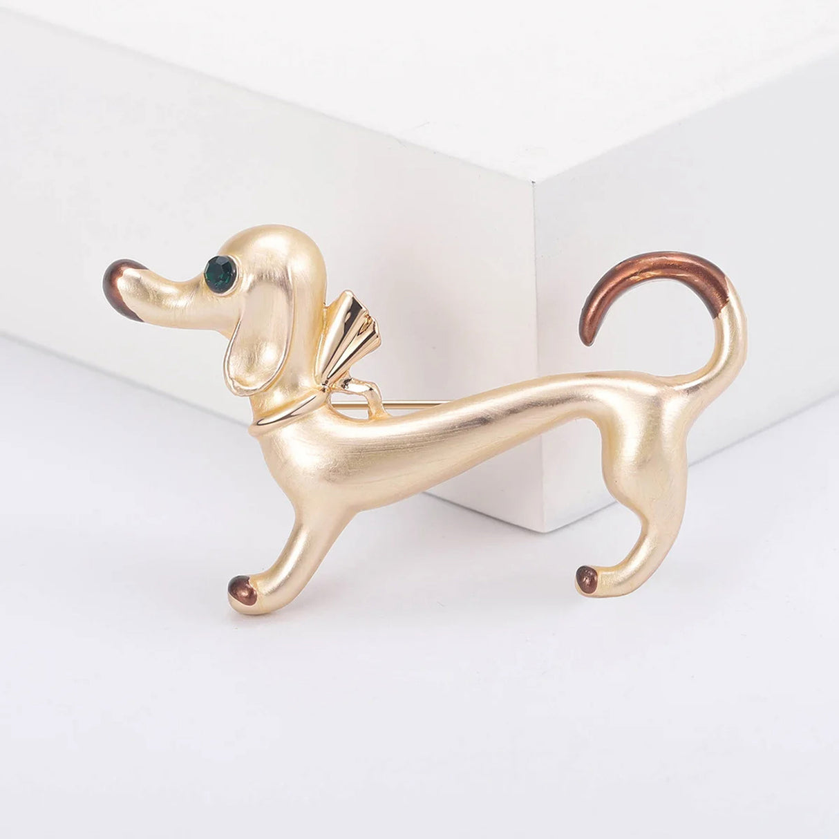Dachshund dog brooch in polished gold-tone enamel with brown accents, green faceted eye, and gold-tone bow, upright pose on white studio backdrop.