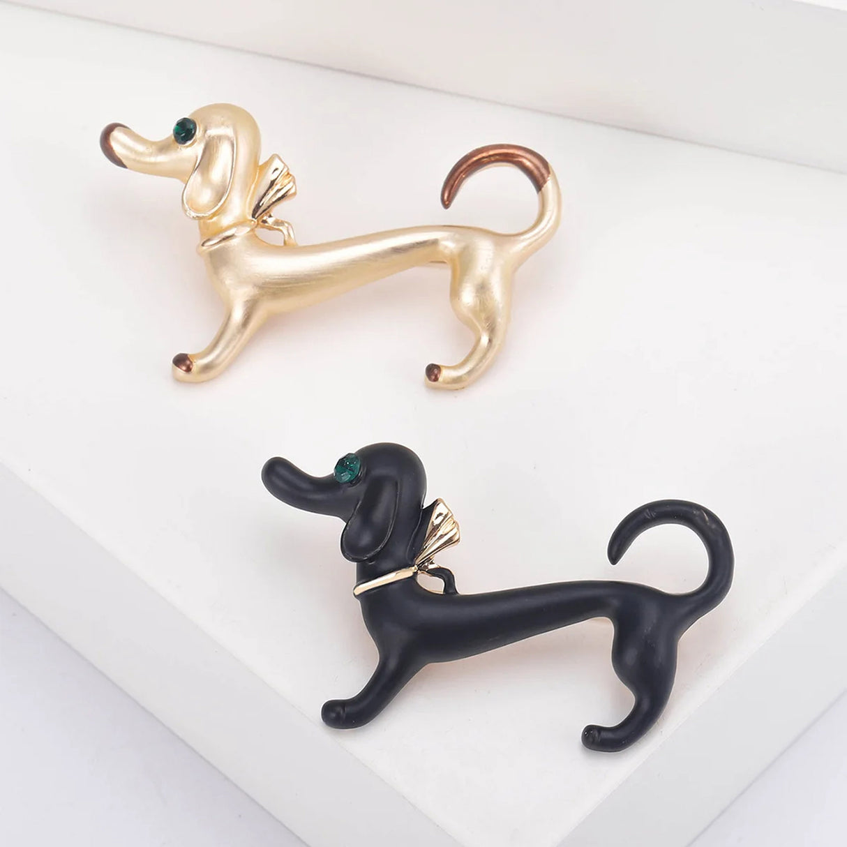 Pair of dachshund dog brooches, one in black and one in gold-tone with brown tips, both with green eyes and gold-tone bows, angled view on white studio background.