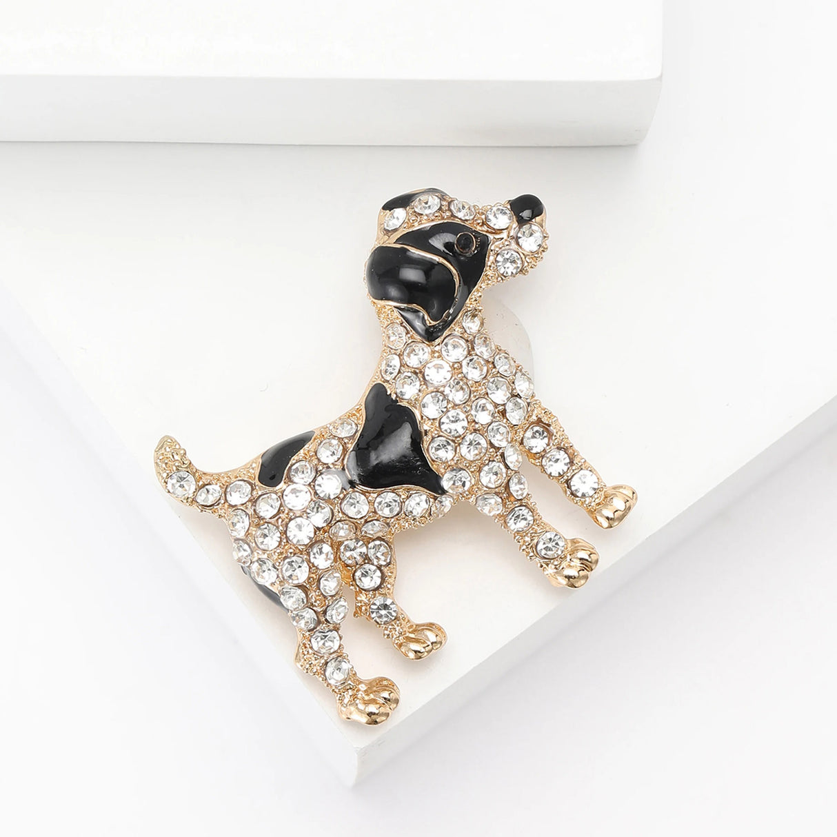 Dog brooch with gold-tone base metal, black enamel patches, and clear faceted stones. Angled top-down view on white studio surface.