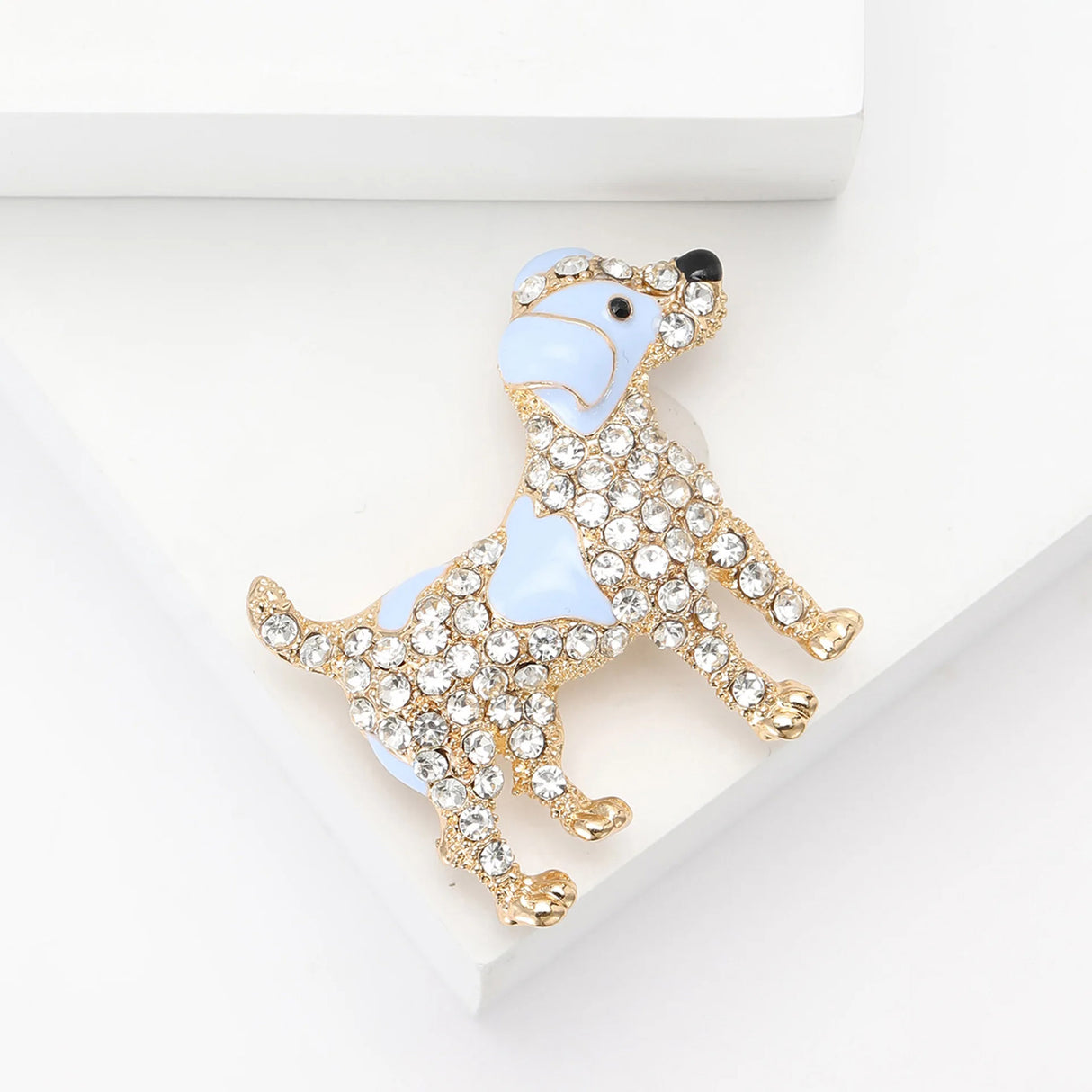 Dog brooch with light blue enamel patches, clear faceted stones, and gold-tone outline. Angled bird's eye view on white background in studio.