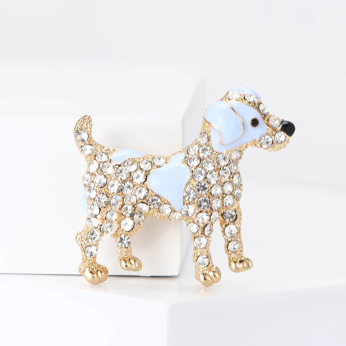 Dog brooch in gold-tone metal finish with light blue enamel accents and clear stones, standing upright on white block.