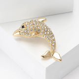 Dolphin brooch with gold-tone finish, clear faceted stones, and black eye detail, flat lay on white blocks.