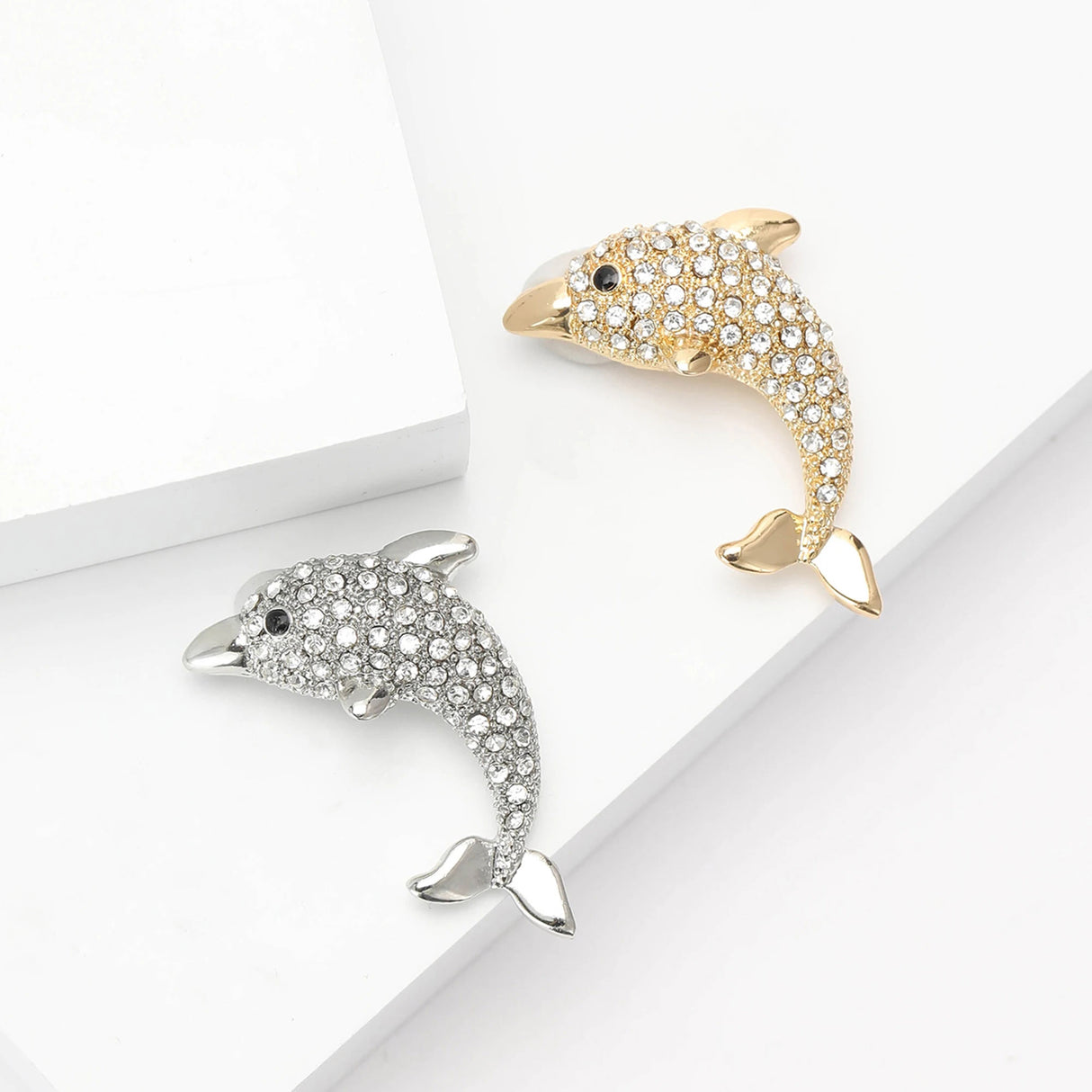 Pair of dolphin brooches with clear faceted stones, one gold-tone and one silver-tone, flat lay on white.