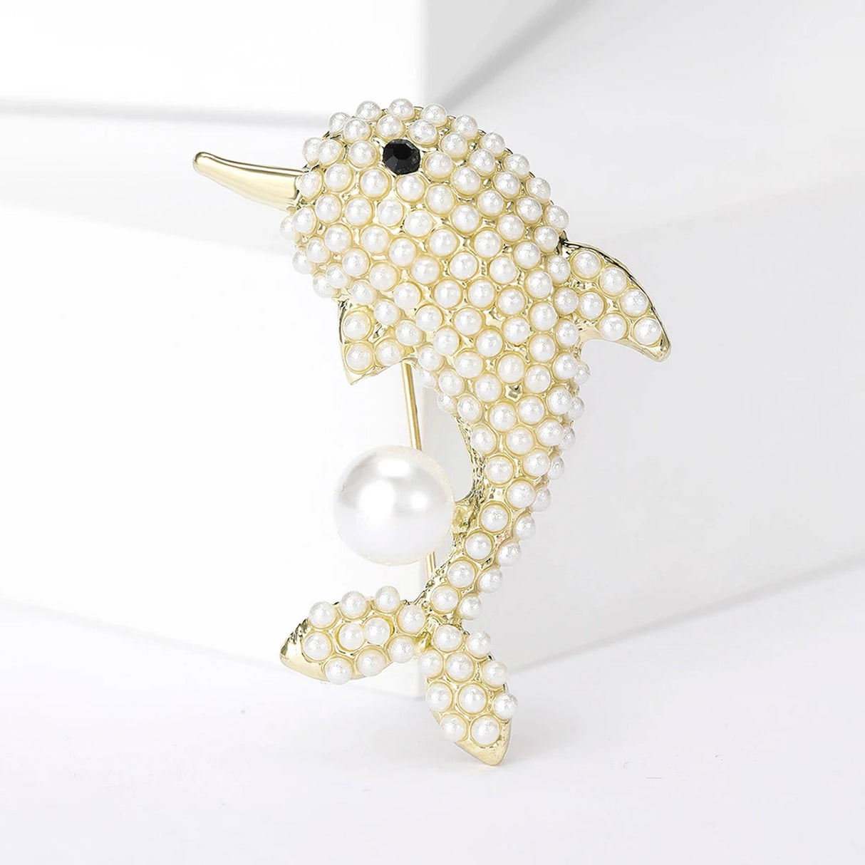 Dolphin brooch with gold-tone base, white round beads, black eye detail, and a single large faux pearl, upright pose on white studio backdrop.