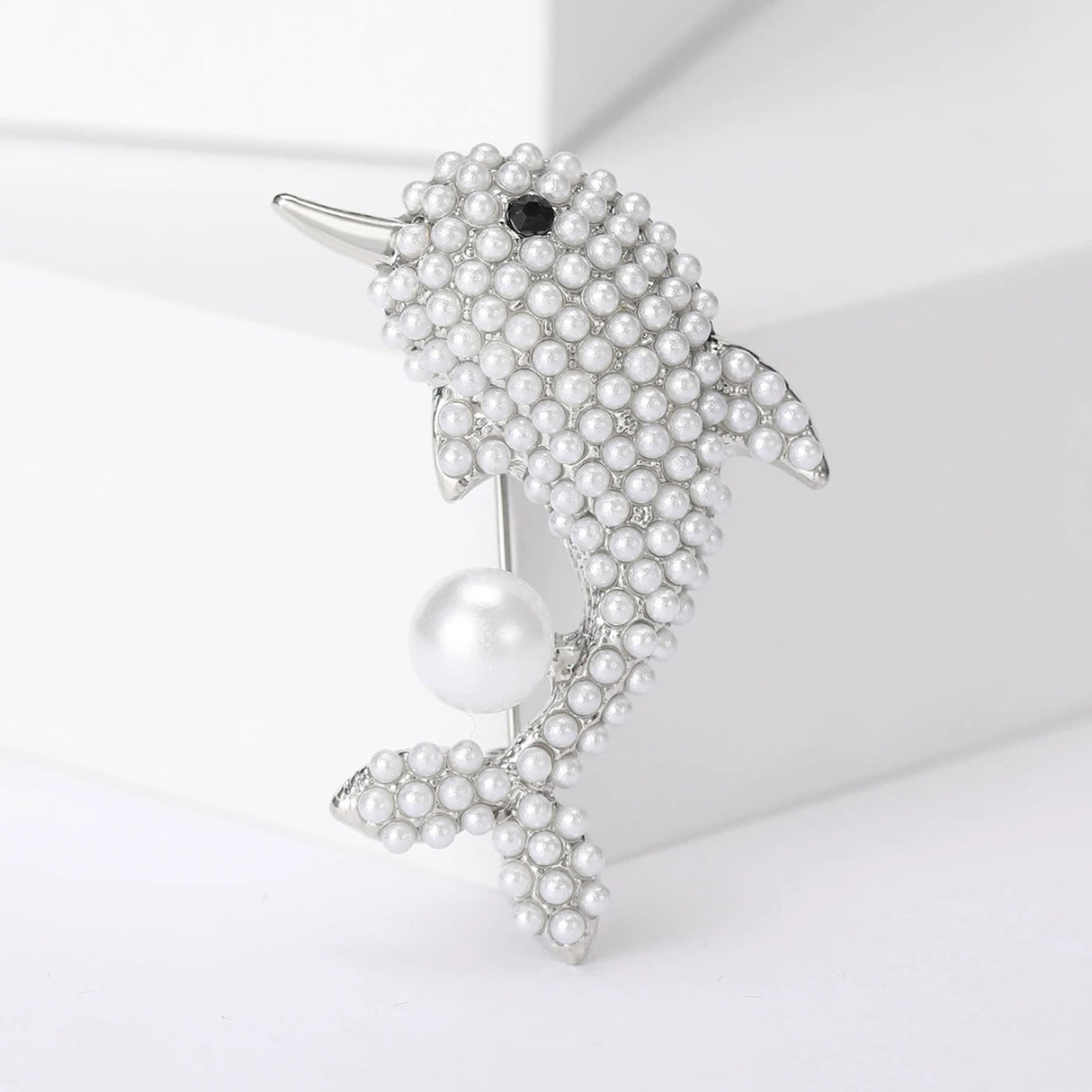 Dolphin brooch with silver-tone metal base, small white round beads, black eye accent, and a large imitation pearl, upright pose on white studio background.