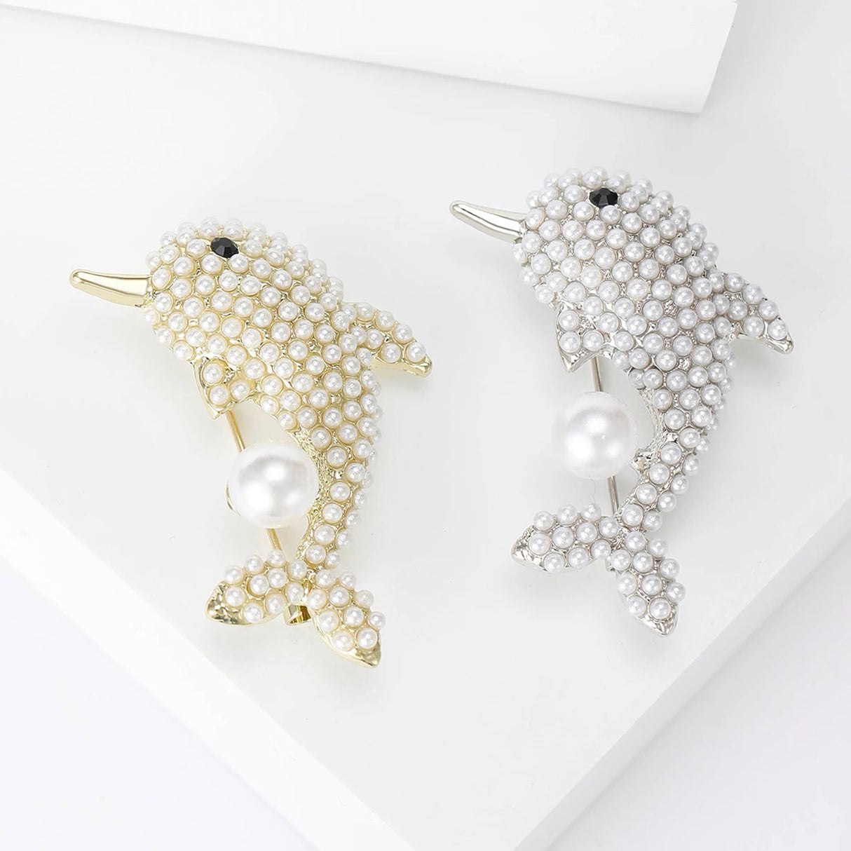 Pair of dolphin brooches with gold-tone and silver-tone bases, covered in small white beads, and large hanging faux pearl, top-down view on white.