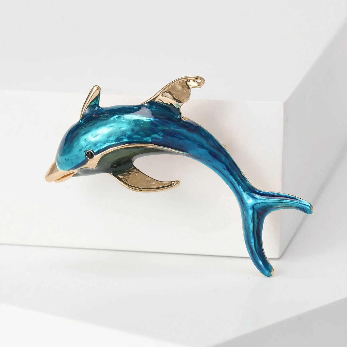 Dolphin brooch with blue enamel finish and gold-tone accents, angled view on white block surface in studio.