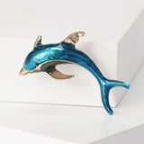 Dolphin brooch with blue enamel finish and gold-tone accents, angled view on white block surface in studio.
