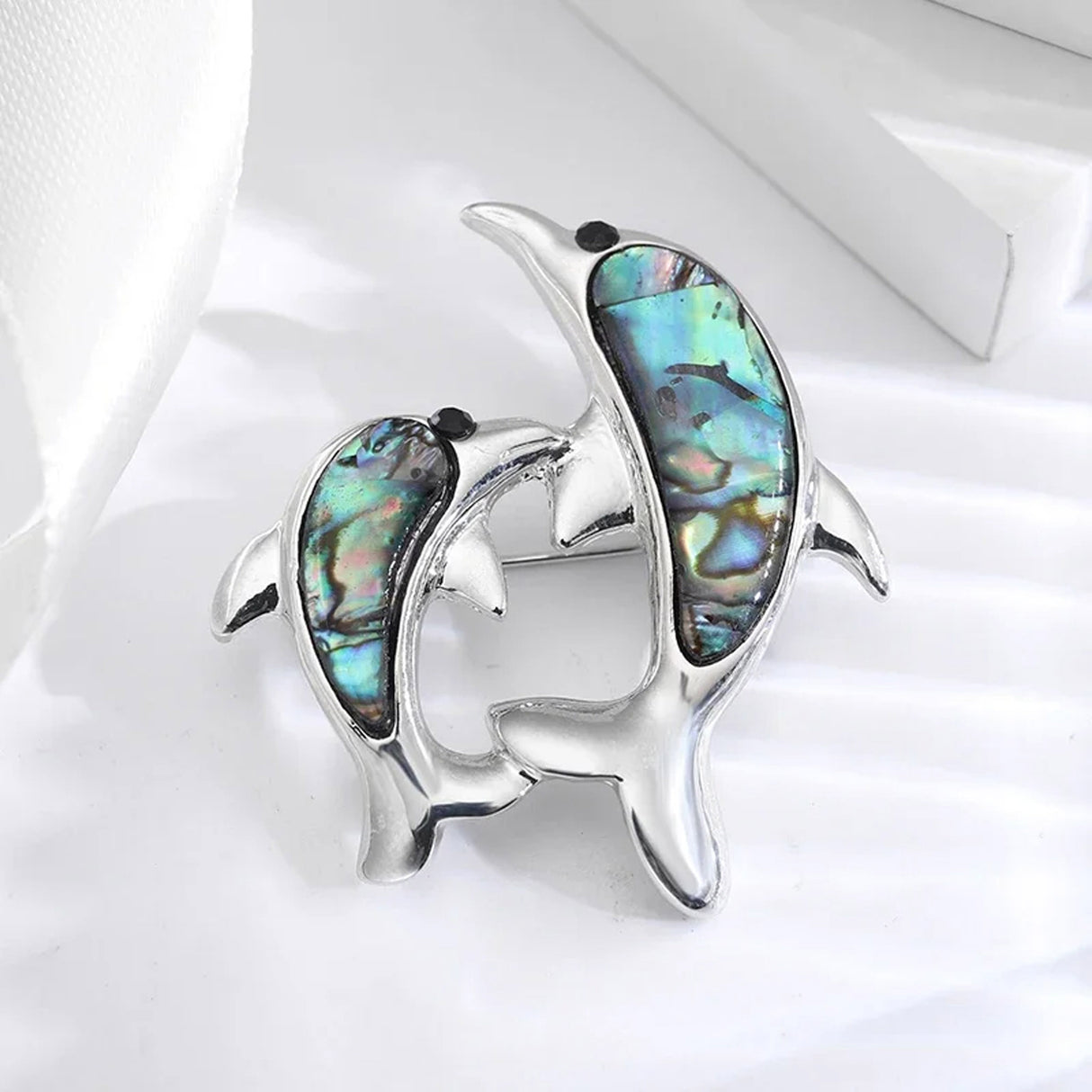 Dolphin brooch with silver-tone metal, multicolor iridescent inlays resembling faux abalone shell, and black bead eyes, angled view on white block.