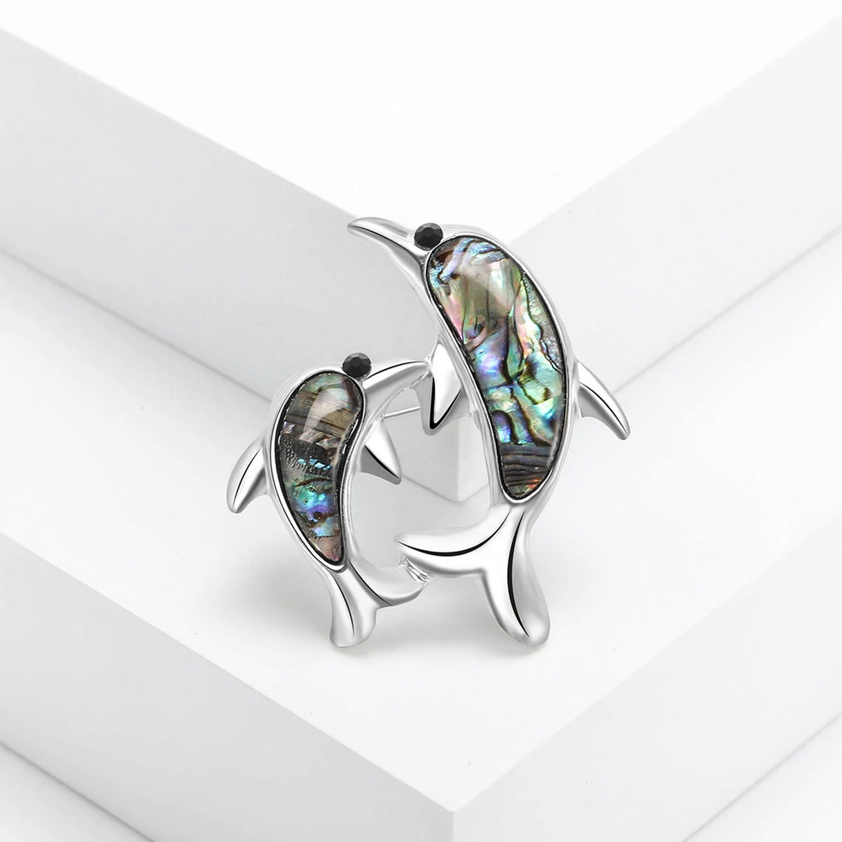 Brooch featuring two silver-tone dolphins with multicolor iridescent inlay evoking imitation abalone shell and black eyes, displayed on studio white surface.