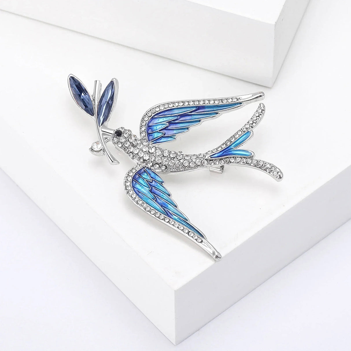 Dove brooch featuring blue enamel accents on wings, faceted stone leaf in beak, and clear stone pave along body and wings. Angled top view on white background.