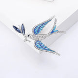 Dove brooch with blue-toned enamel wings, dark blue leaves, and clear faceted stones, top view on white platform.