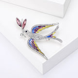 Dove brooch with yellow, purple, and blue-toned wings, pink marquise leaf detail, and white pave across the body. Angled top view on white block.