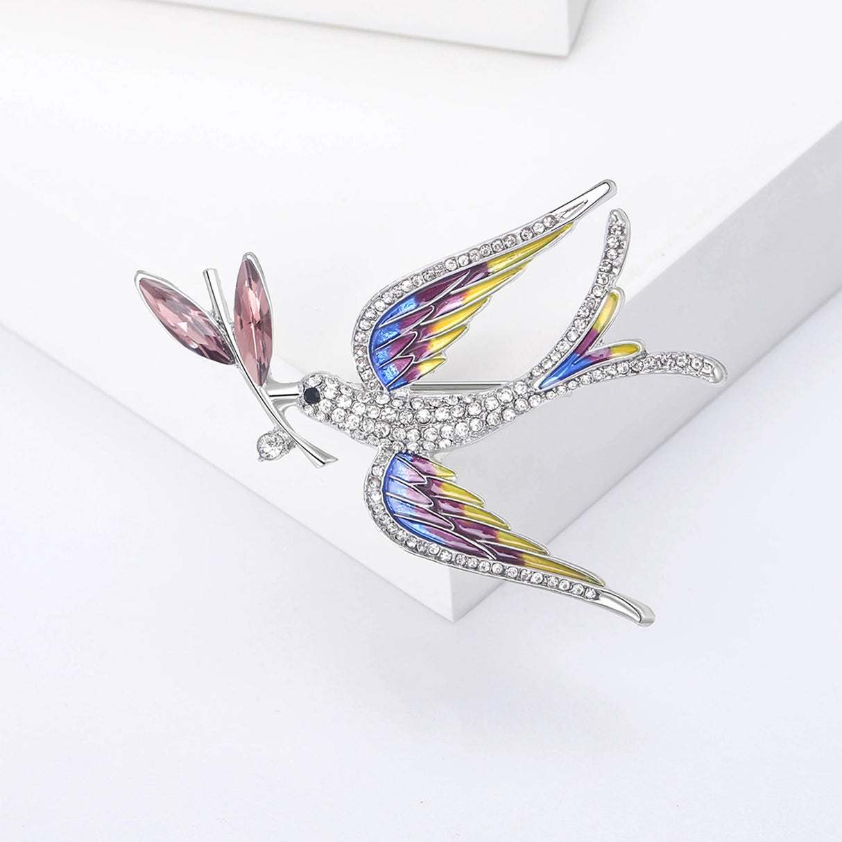 Multicolor dove brooch with layered wing colors in yellow, pink, and blue, pink leaf elements, and clear stones, front view on white squares in studio.