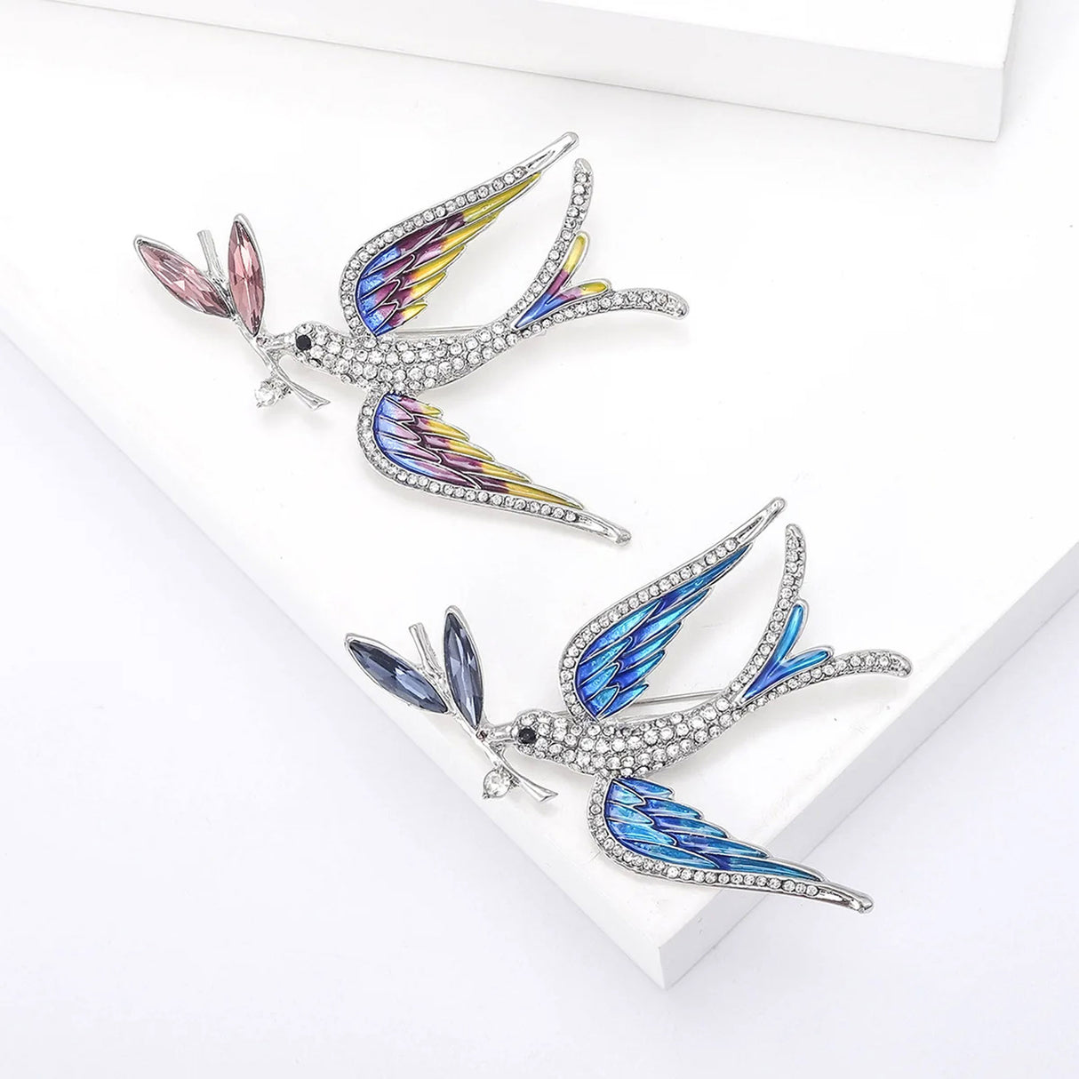 Set of two dove brooches with clear faceted stones, one with blue-toned wings and dark leaves, one with multicolor wings and pink leaf detail, top-down view on white.