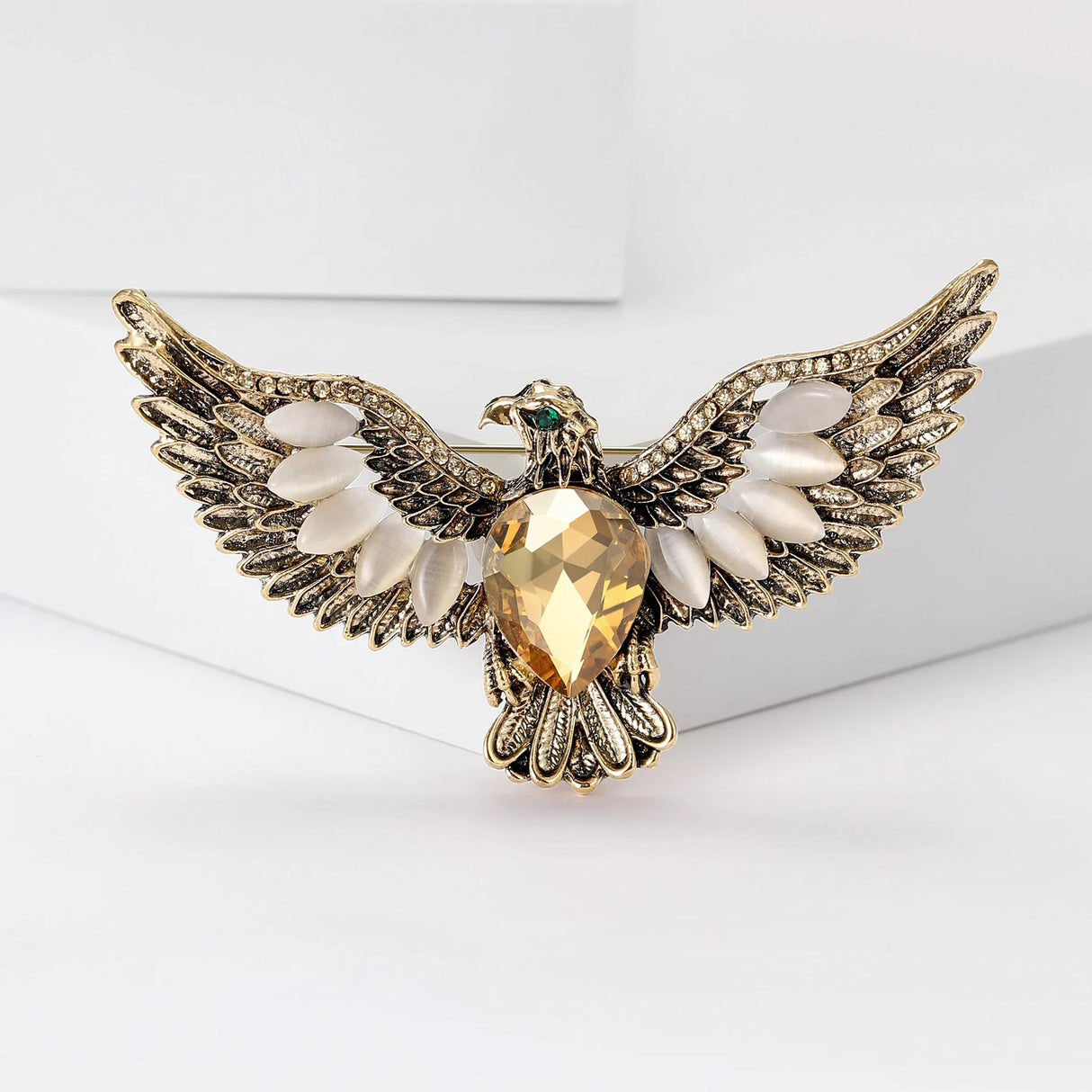 Eagle brooch in gold-tone metal with large yellow faceted stone, white oval accent beads, and green eye detail, upright pose on white platform.