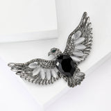 Eagle brooch with silver-tone metal, black faceted center stone, white oval beads, and green eye accent, angled view from top on white surface.