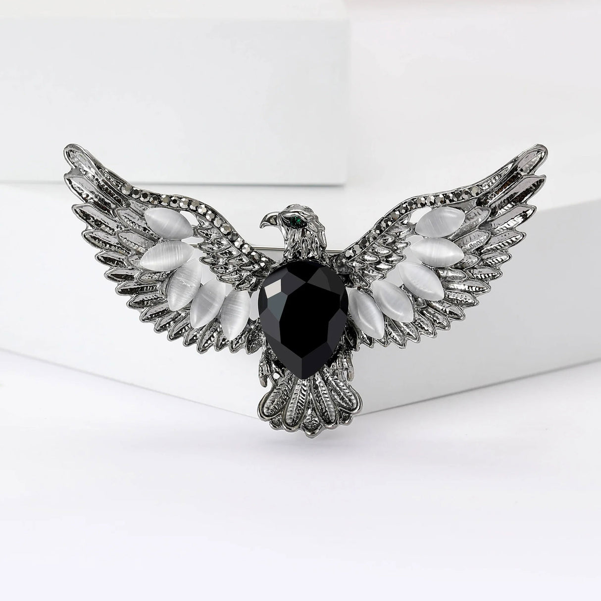 Eagle brooch in silver-tone metal with black faceted center stone, white oval beads, and green eye detail, upright view on white block.