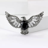 Eagle brooch in silver-tone metal with black faceted center stone, white oval beads, and green eye detail, upright view on white block.