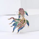 Fish brooch in gold-tone metal with pink and clear stones, blue-green enamel fins, and imitation pearl, upright on white surface.