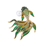Fish brooch with gold-tone metal setting, green and clear faceted stones, green and yellow enamel fins, and white round bead, cutout on white.