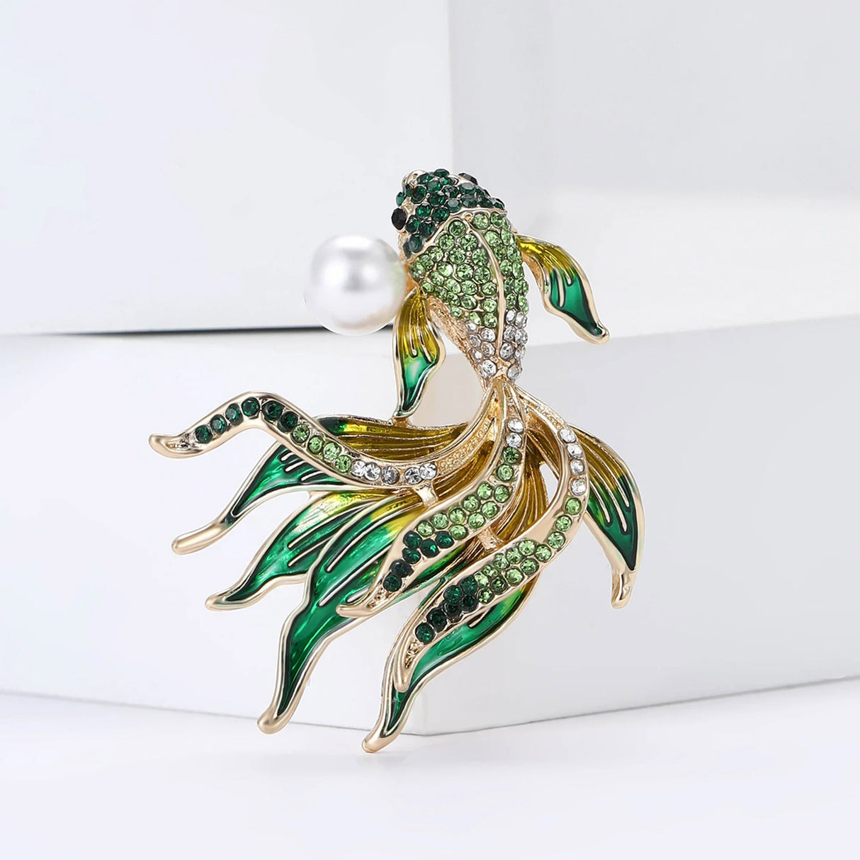 Fish brooch with green faceted stones, yellow and green enamel fins, clear accents, and faux pearl, upright studio setting.