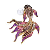 Fish brooch with gold-tone outline, purple faceted stones, pink-toned enamel fins, and white round bead, cutout on white.