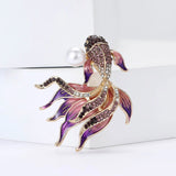 Fish brooch with purple and clear faceted stones, pink-purple enamel fins, and imitation pearl, upright on white block.