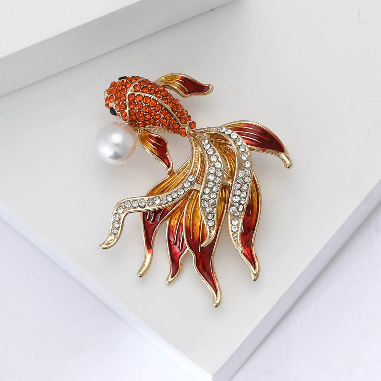 Fish brooch with orange faceted stones, red and yellow enamel fins, clear accents, and faux pearl, flat lay on white.