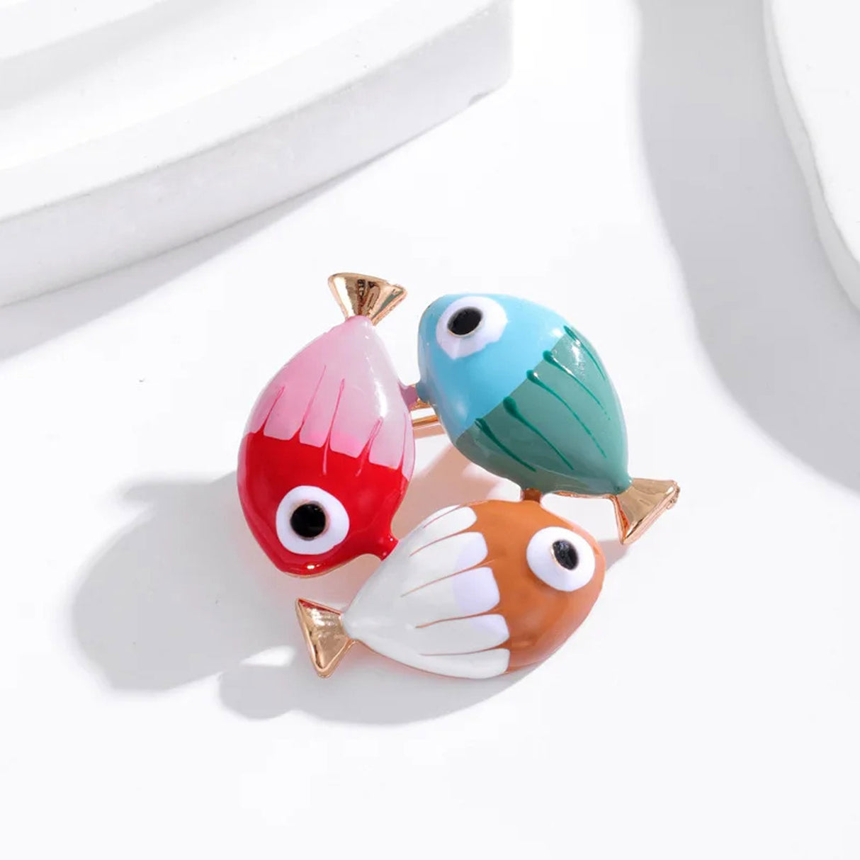 Fish brooch with red and pink, blue and green, and white and orange enamel finish, gold-tone tails, studio flat lay on white with shadows.
