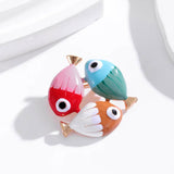 Fish brooch with red and pink, blue and green, and white and orange enamel finish, gold-tone tails, studio flat lay on white with shadows.