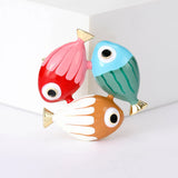 Fish brooch with red-pink, blue-green, and orange-white enamel details and gold-tone accents, upright view on white block in studio.