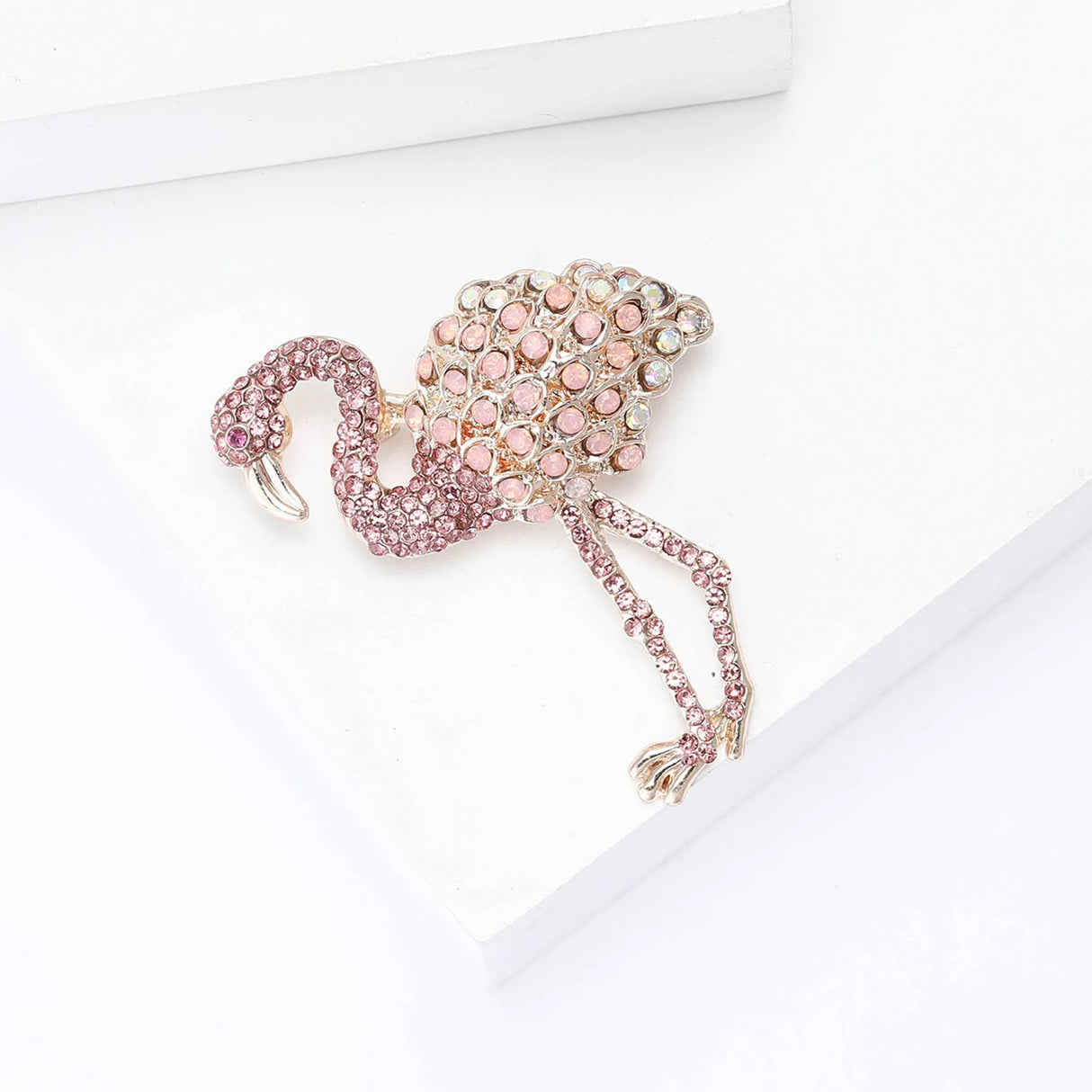 Flamingo brooch with pink stones, round iridescent pink accents, and gold-tone metallic legs. Top-down view on white blocks.