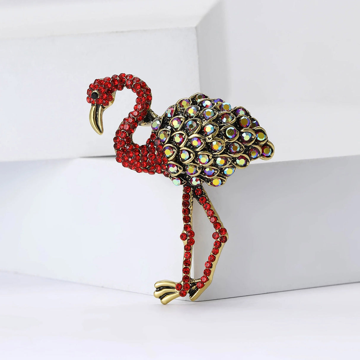 Flamingo brooch with red-tone stones and multicolor reflective teardrop accents, standing view on white blocks.