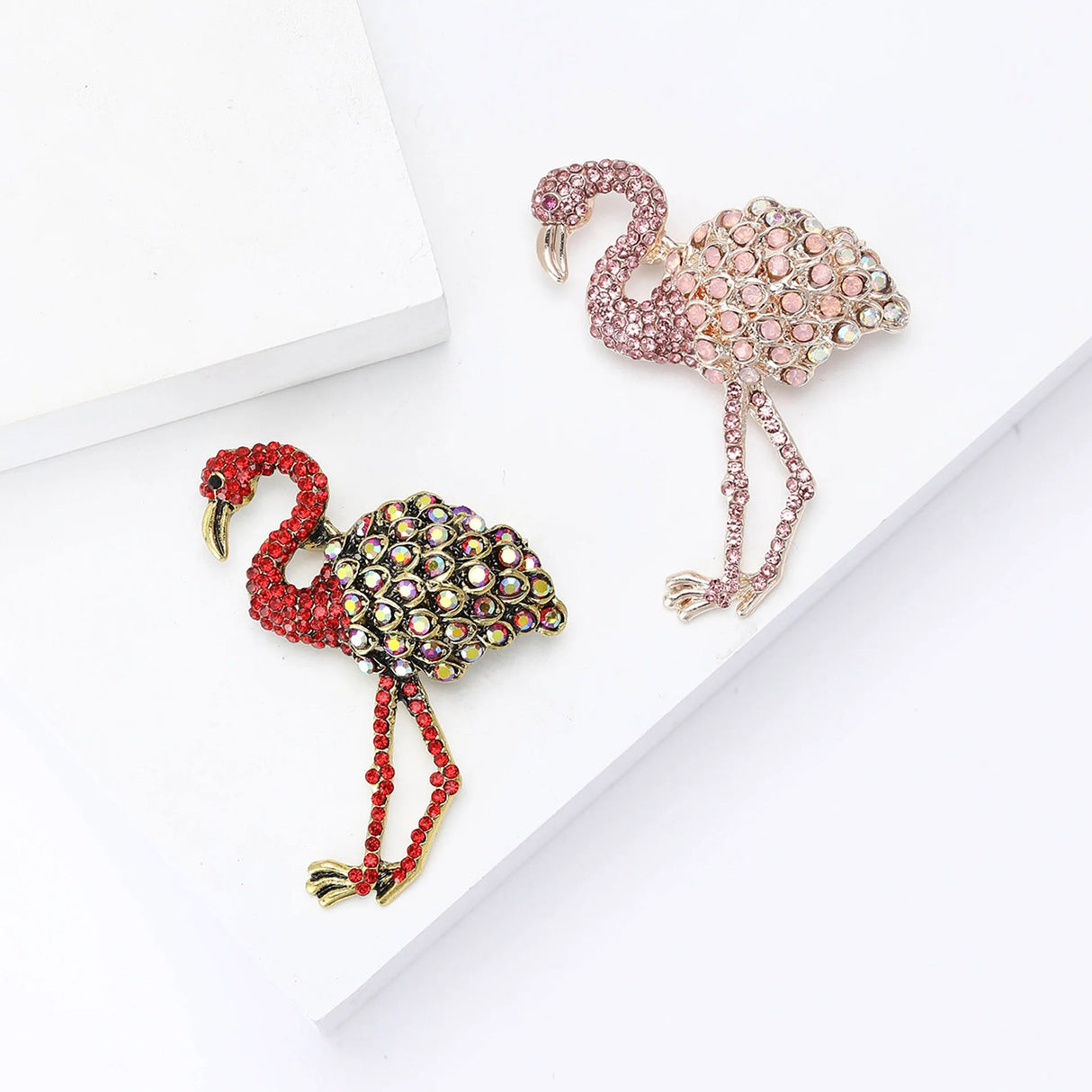 Set of two flamingo brooches with red and pink-tone stones and multicolor accents, flat lay on white surface.