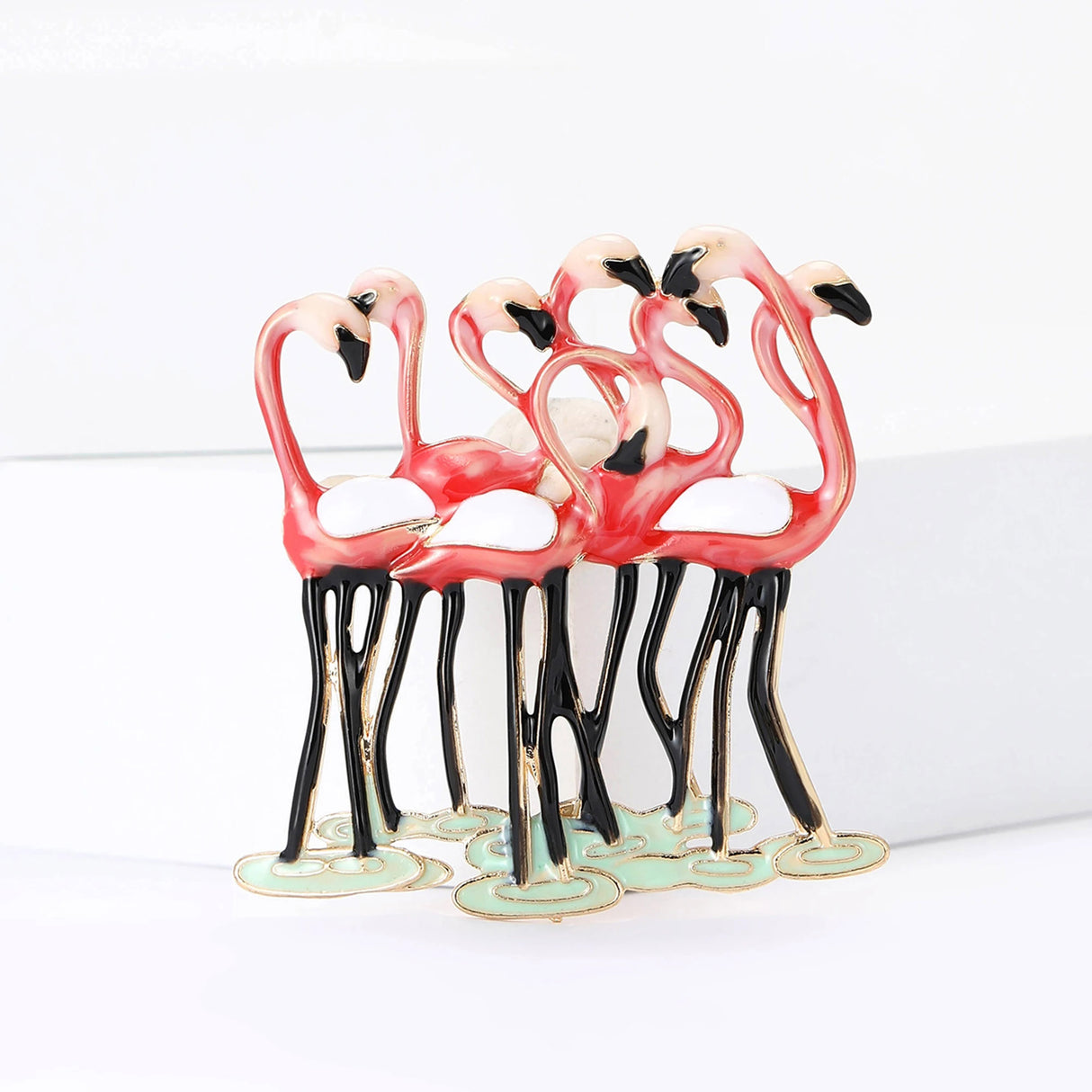 Flamingo brooch showing group of pink birds with black legs, white wings, and green base, displayed on white block.