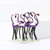 Flamingo brooch with group of purple birds, featuring black legs and white wings, shown on white platform.