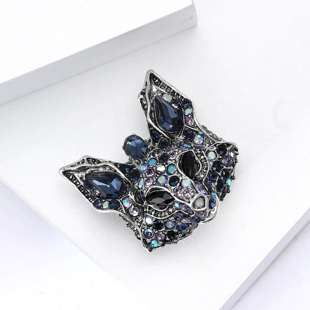 Fox brooch in silver-tone metal with stones in multiple shades of blue and black eyes, angled view on white block.