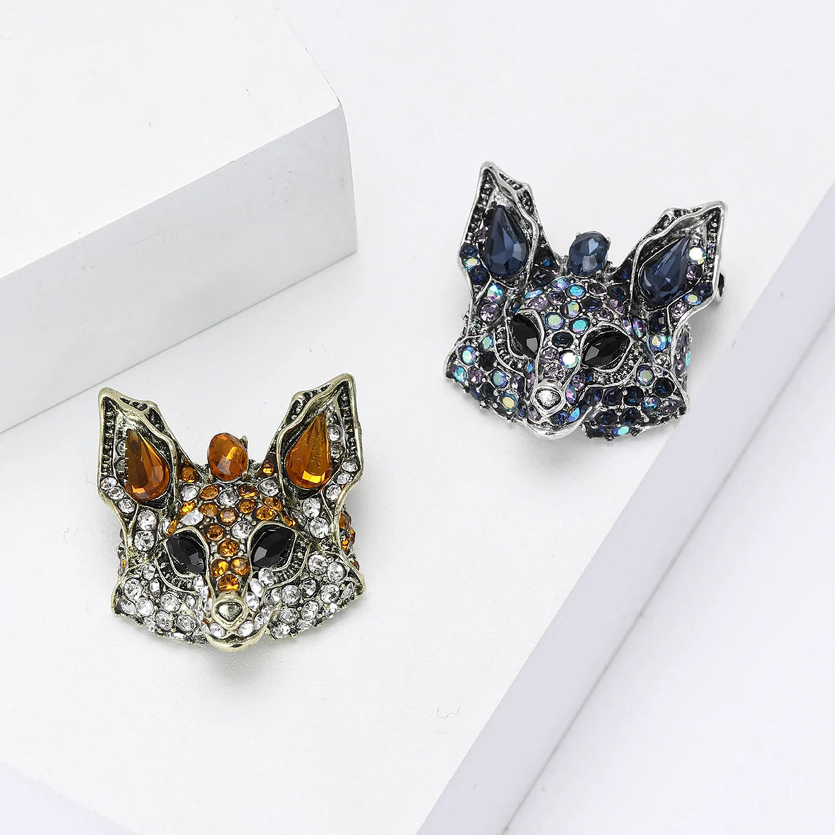 Two fox brooches, one with orange and clear stones and the other with blue and iridescent accents, flat lay on white surface.